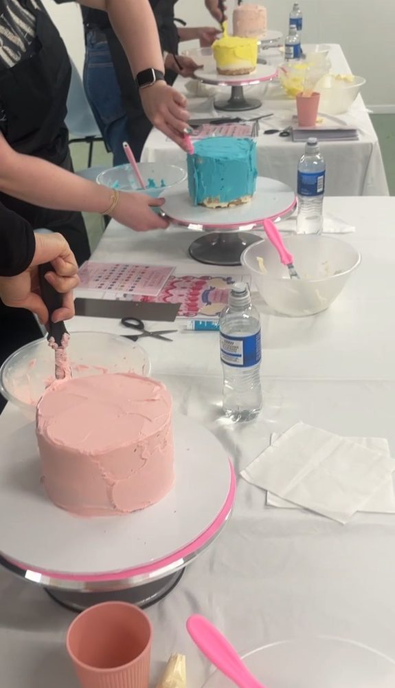 Class table with pink, blue and yellow cakes being decorated